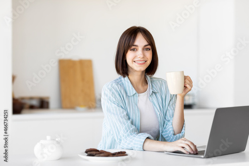 Happy millennial woman sitting in kitchen with laptop computer and drinking hot coffee  smiling at camera