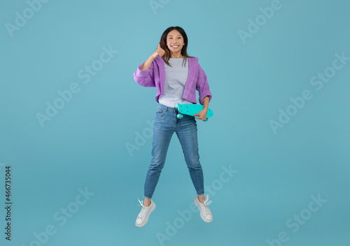 I like teenage culture. Joyful asian lady with skateboard jumping and showing thumb up over blue background