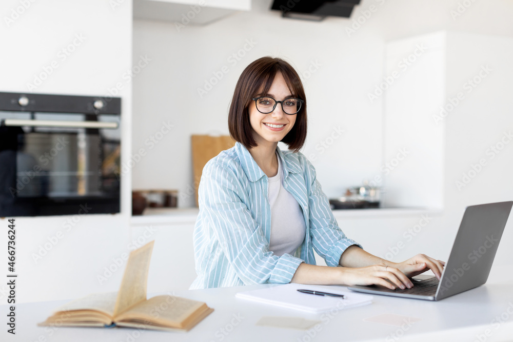 Happy young freelancer working on laptop in kitchen interior, sitting at table and smiling at camera Happy young freelancer working on laptop in kitchen interior, sitting at table and smiling at camera
