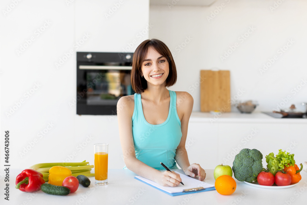 Happy fitness lady in sportswear writing daily ration diet or healthy recipe, standing in kitchen Happy fitness lady in sportswear writing daily ration diet or healthy recipe, standing in kitchen