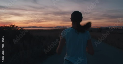 Girl running against the backdrop of an epically beautiful pink sunset with house lights reflecting in a mountain lake. Brunette hair swaying from side to side while running. Female in white t-skirt. photo