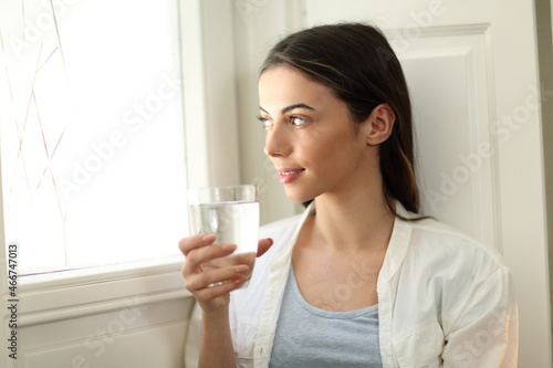 Woman holding water glass lokking through window at home