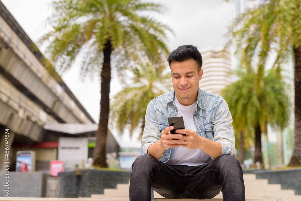Portrait of handsome young man sitting outdoors in city during summer while using mobile phone Portrait of handsome young man sitting outdoors in city during summer while using mobile phone