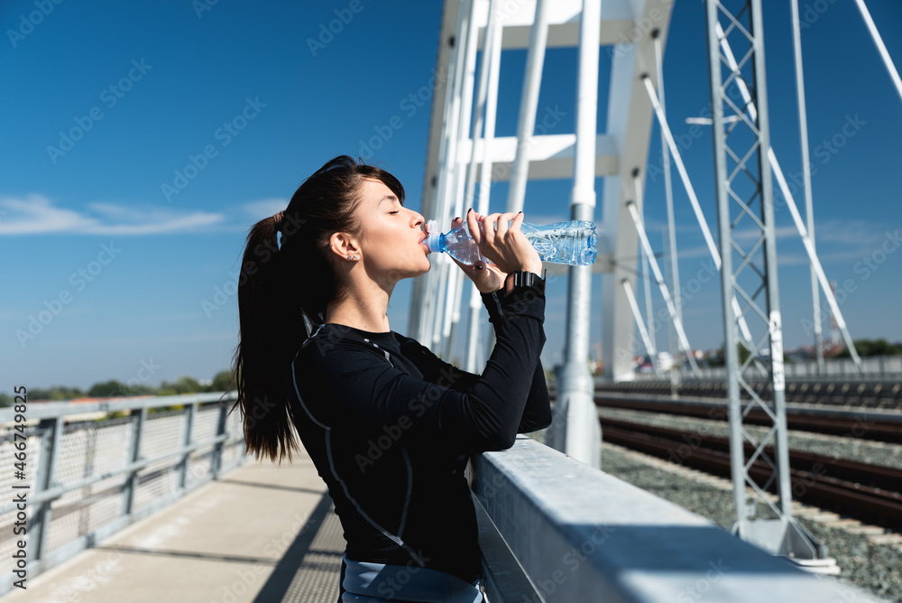 Young happy self loved sporty fitness runner woman standing and taking a break drinking bottle of water after hard running and jogging exercise for healthy life and clear mind preparing for marathon