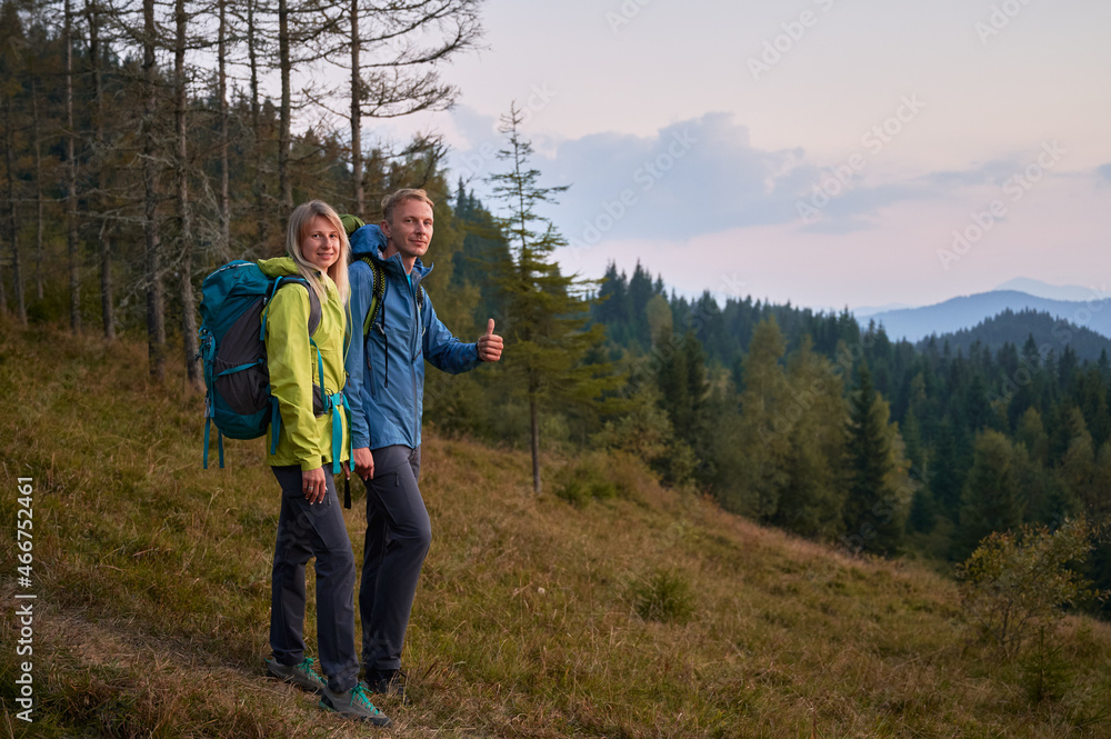 Smiling husband and wife with backpacks standing on grassy downhill trail against the backdrop of mountain forests. Positive male while trekking adventure showing cool gesture.