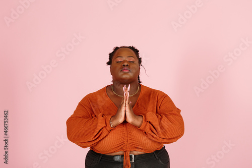 Hopeful young black plus size body positive woman with dredlocks in orange top prays with closed eyes on pink background in studio closeup