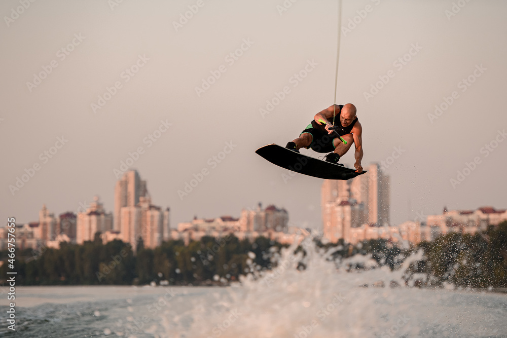 active man holds rope and skilfully jumping high in air on wakeboard over splashing water.