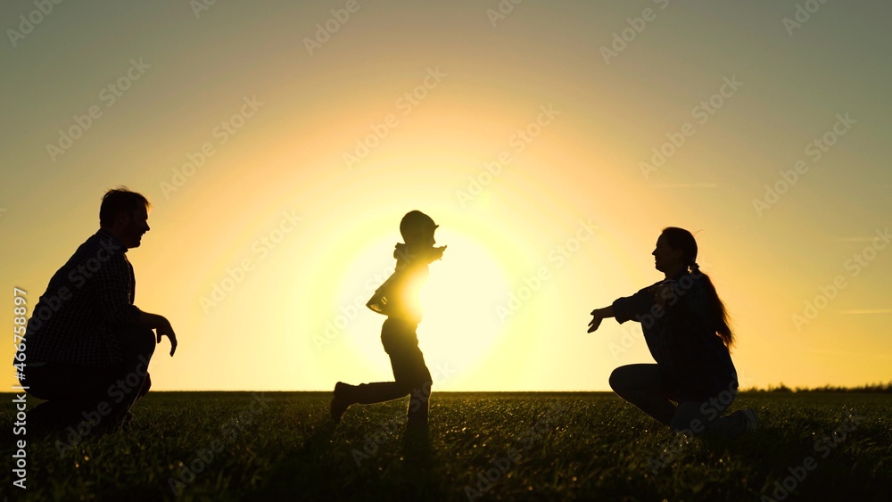Happy family is playing in park at sunset. Daughter runs from dad to mom in sun. Happy family and childhood concept. Family weekend in the spring on grass. Walk with small kid in nature.