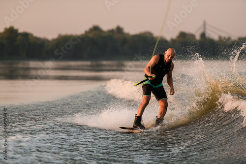 active man holds cable and energetically rides down on splashing wave on wakeboard.