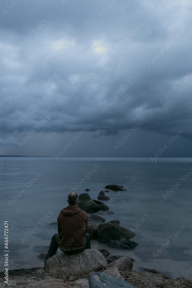 Fototapeta premium Mann sitzt alleine auf einem Stein am Meer mit dunklen Regenwolken am Himmel.