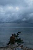 Mann sitzt alleine auf einem Stein am Meer mit dunklen Regenwolken am Himmel.