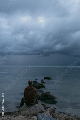 Mann sitzt alleine auf einem Stein am Meer mit dunklen Regenwolken am Himmel. © Lars Gieger