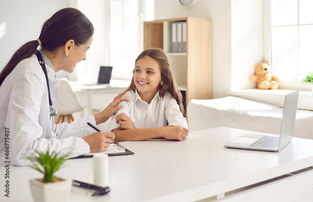 Friendly and caring female doctor encourages little girl before medical examination. Pediatrician or nurse sits at table with clipboard and touches child's shoulder giving her support. Trust concept.