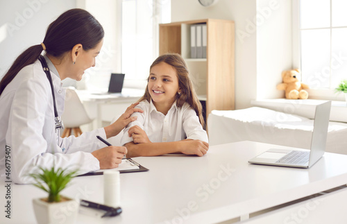 Friendly and caring female doctor encourages little girl before medical examination. Pediatrician or nurse sits at table with clipboard and touches child s shoulder giving her support. Trust concept.