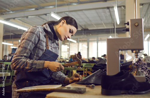 Female worker using professional tools while making new leather boots. Beautiful young woman working with hammer and nail at shoe factory workshop. Manufacturing industry, footwear production concept