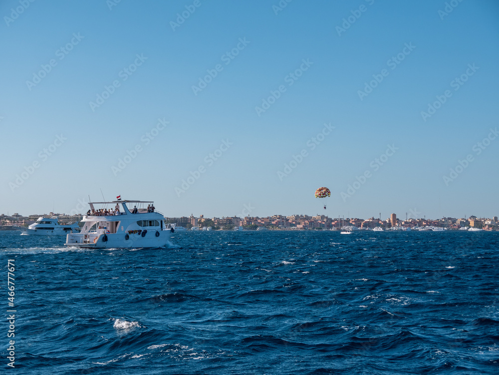 A boat with tourists sails along the coast with hotels. The boat pulls a multi-colored parachute with tourists.