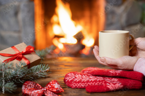A Christmas gift, women's hands holding a mug of hot mulled wine, red knitted mittens and fir decorations on the background of a burning fire in the fireplace. Soft focus. photo