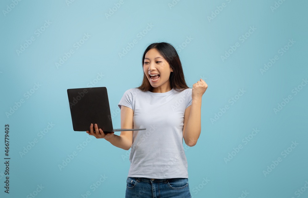 Euphoric asian lady celebrating success with laptop, raising clenched fist while standing over blue studio background Euphoric asian lady celebrating success with laptop, raising clenched fist while standing over blue studio background