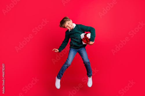 Photo of funny cute schoolboy dressed green sweater jumping high holding gift box smiling isolated red color background