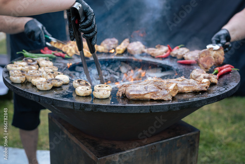 Several hands use tongs to turn mouth-watering pork steaks, sausages and open-air grilled kebabs. City picnic.