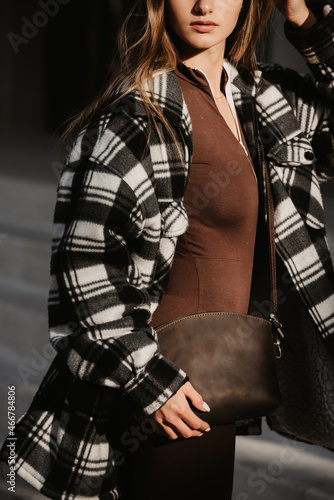 Outdoor fashion portrait of young beautiful fashionable girl wearing trendy checkered shirt, solid long sleeve bodycon one piece jumpsuits and small brown color small bag, posing in street