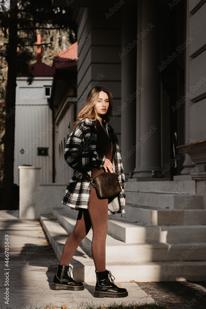 Naklejka premium Outdoor fashion portrait of young beautiful fashionable girl wearing trendy checkered shirt, solid long sleeve bodycon one piece jumpsuits and small brown color small bag, posing in street