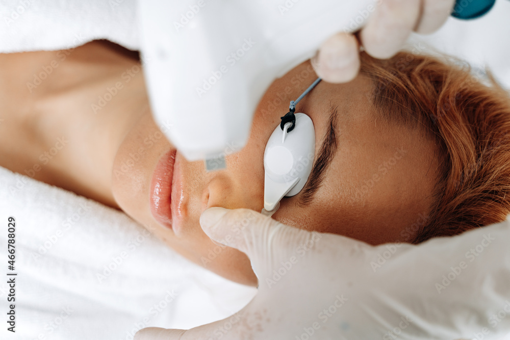 Naklejka premium Close up cosmetologist arm making depilation of face for female client. Ginger woman laying at the clinic during her treatment