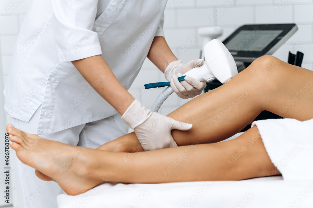 Aesthetic body treatment. Side on cropped head portrait of young slender relaxed woman in white bathrobe having hair removal procedure of legs in knee zone Aesthetic body treatment. Side on cropped head portrait of young slender relaxed woman in white bathrobe having hair removal procedure of legs in knee zone