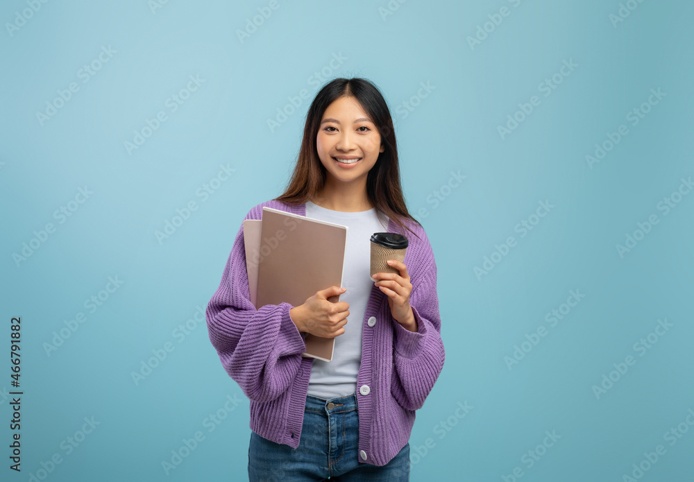 Excited asian female student holding notebooks and coffee to go, having break while studying, blue background Excited asian female student holding notebooks and coffee to go, having break while studying, blue background