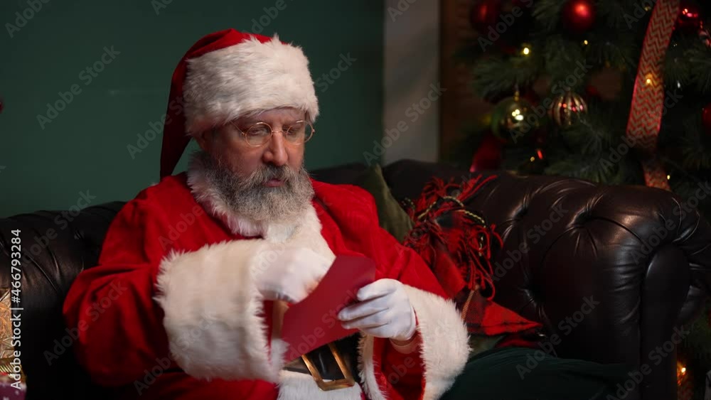Santa Claus reads letters with delight and a smile on his face. Old man in a red suit and a hat sits on a sofa in a decorated room near a glowing Christmas tree and gifts. Close up. Slow motion. Santa Claus reads letters with delight and a smile on his face. Old man in a red suit and a hat sits on a sofa in a decorated room near a glowing Christmas tree and gifts. Close up. Slow motion.