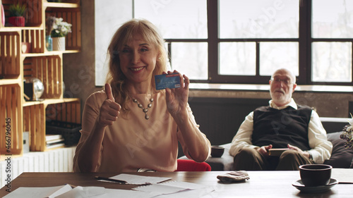 Senior caucasian woman holding credit card and showing thumbup sitting at table at home.