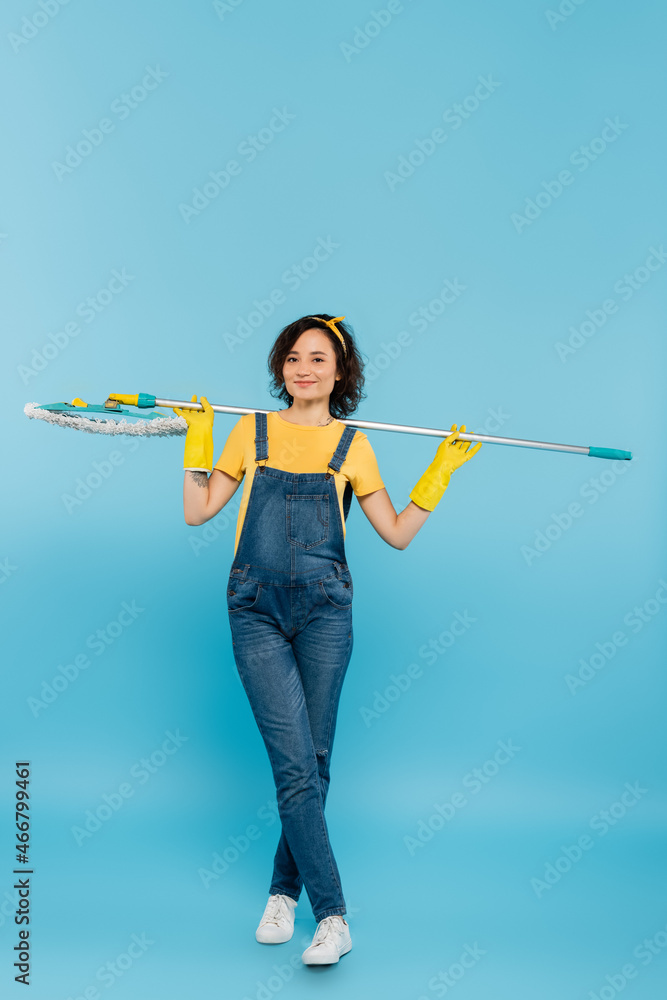 happy housewife in yellow rubber gloves and denim overalls posing with mop on blue. happy housewife in yellow rubber gloves and denim overalls posing with mop on blue.