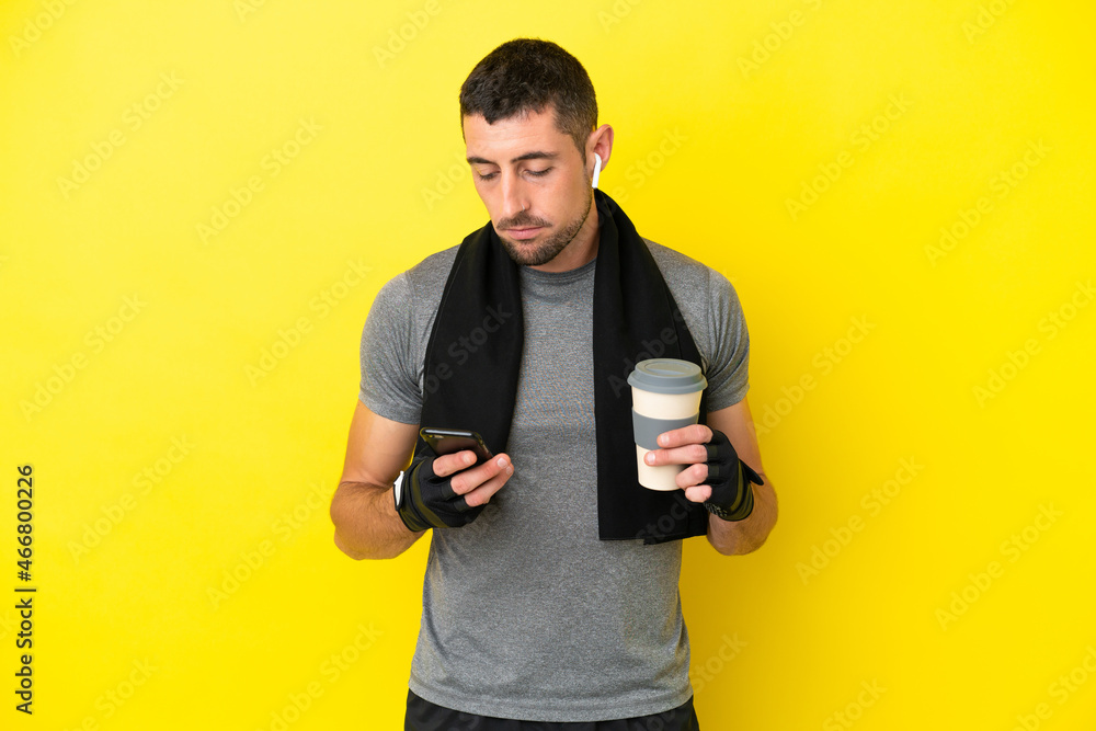 Young sport caucasian man isolated on yellow background holding coffee to take away and a mobile Young sport caucasian man isolated on yellow background holding coffee to take away and a mobile