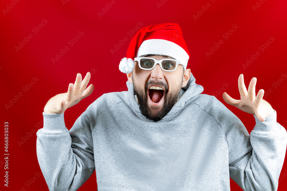 Naklejka premium on a red Christmas background, a man in a Santa Claus hat and with glasses for watching movies, a man screams from what he saw
