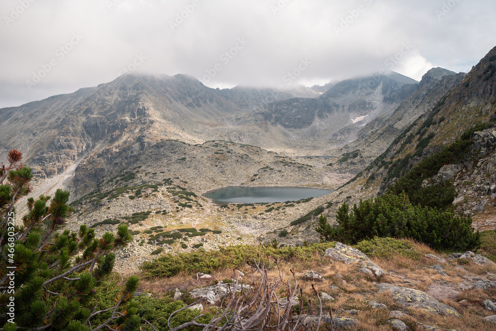 Stunning view towards Musala summit on Rila mountain, over foreground plants, glacier lake and rocky peaks covered in clouds and fog