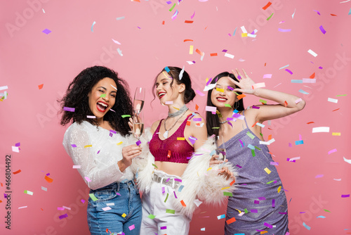 Positive interracial women holding champagne near confetti on pink background
