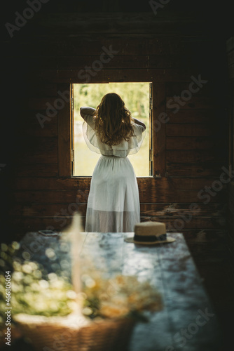 beautiful girl with red hair stands near a wooden window in an old wooden house in a white dress in the foreground a table on which a basket with flowers and a straw hat on a sunny day, rear view the 