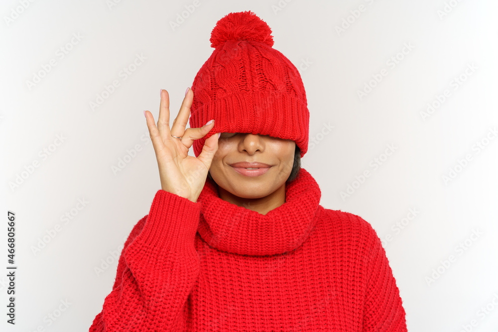 Playful african millennial woman has fun wearing winter hat and sweater covering eyes under cap. Joyful young woman in red woolen knitted winter clothes over studio wall. Wintertime fashion concept Playful african millennial woman has fun wearing winter hat and sweater covering eyes under cap. Joyful young woman in red woolen knitted winter clothes over studio wall. Wintertime fashion concept