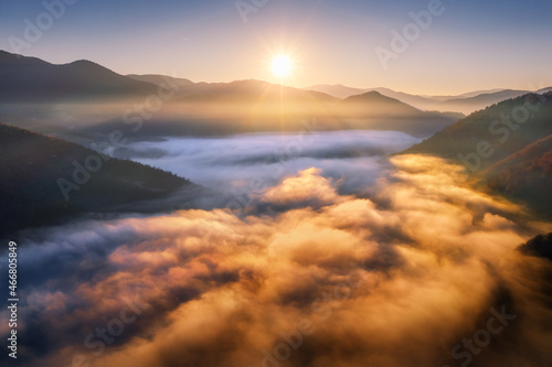 Mountains in low clouds at sunrise in autumn. Aerial view of mountain peak in fog in fall. Beautiful landscape with rocks, forest, sun, colorful sky. Top view of mountain valley in clouds. Foggy hills