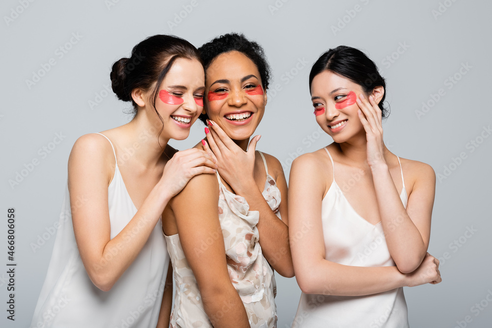 Cheerful multicultural women in silk dresses and eye patches isolated on grey Cheerful multicultural women in silk dresses and eye patches isolated on grey