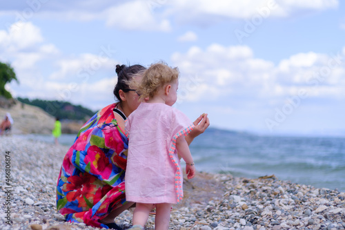 Mother and little girl squat down or crouch at beach, show and teach to throw stone or rock to her daugther, family holiday concept idea with copy space