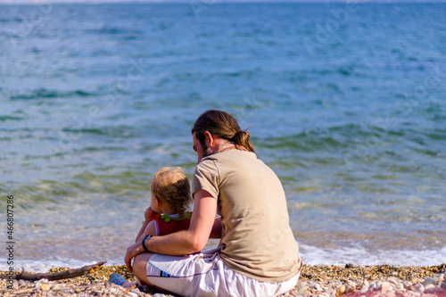 Blonde little girl and young man sit and look at sea or ocean at beach, holiday concept idea