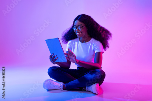 Full length of young black lady using tablet pc, studying or working remotely, having online conference in neon light