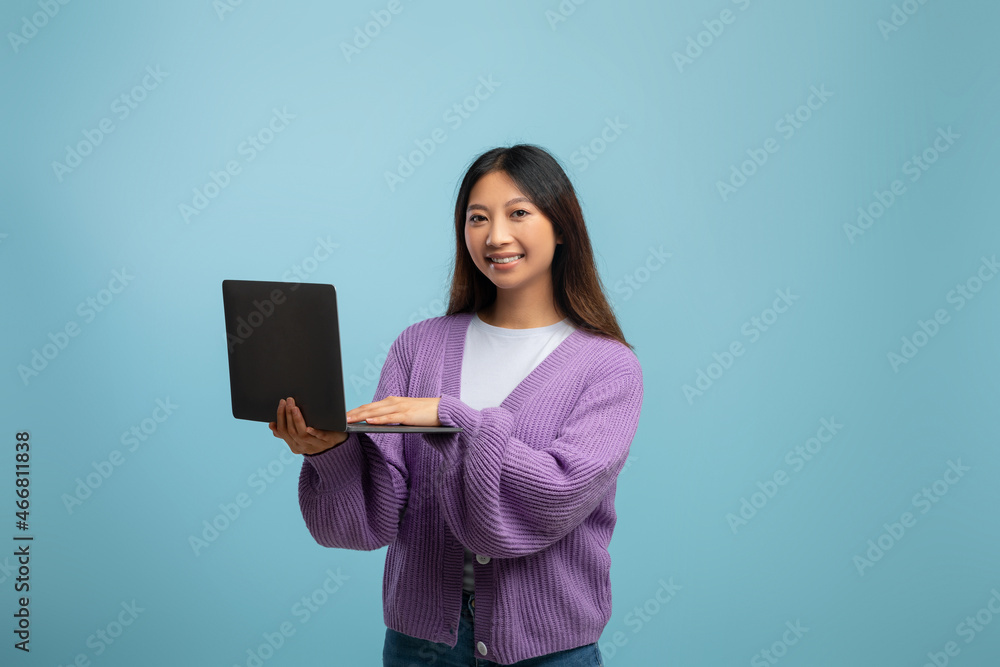 Portrait of happy young asian lady holding and using laptop over blue studio background, smiling at camera Portrait of happy young asian lady holding and using laptop over blue studio background, smiling at camera