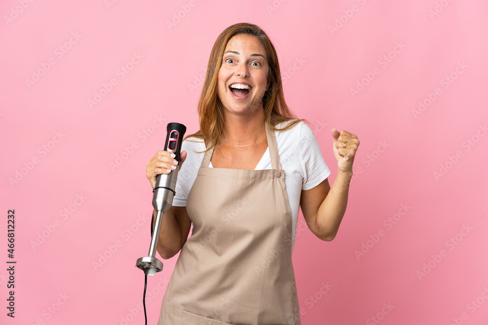 Middle age woman using hand blender isolated on pink background celebrating a victory in winner position Middle age woman using hand blender isolated on pink background celebrating a victory in winner position
