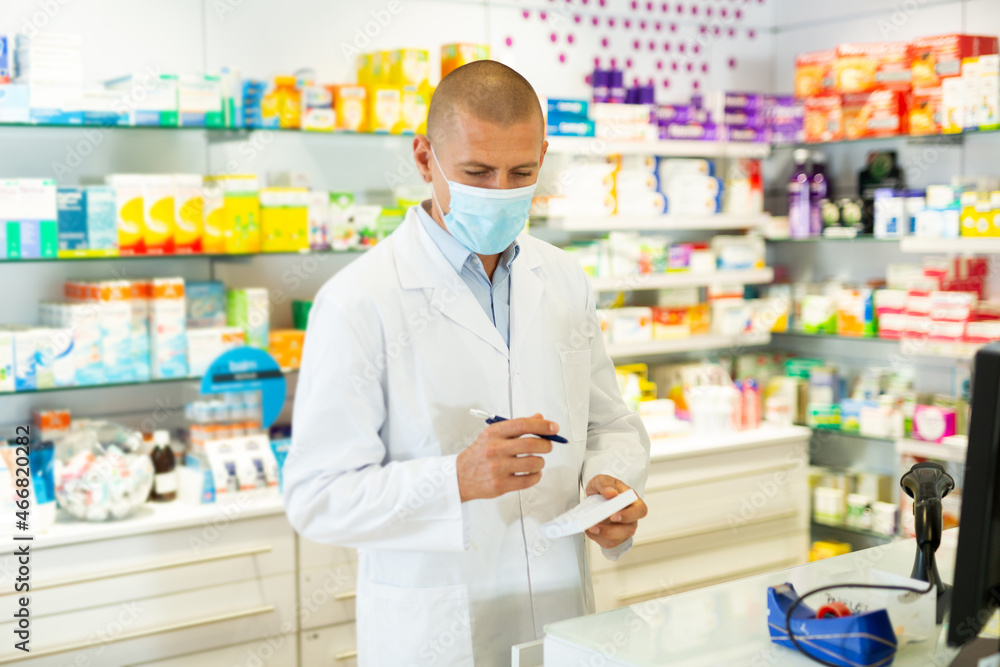 Caucasian male pharmacist in lab coat and face mask standing at counter in chemists shop and writing recipe.