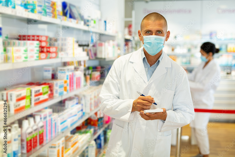 Portrait of an male pharmacist in protective mask, working in pharmacy during the pandemic; standing in trading floor and makes important notes