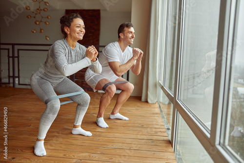 Cheerful sporty man and woman are doing squats with resistance bands before large window indoors
