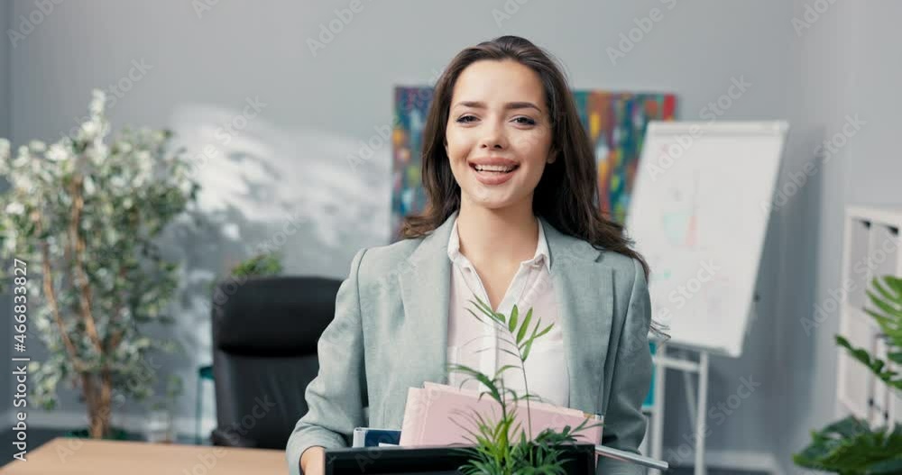 Attractive beautiful smiling brunette in shirt stands in front of desk in company office in hands holds box with packed things, promotion in position, coming to new job in office, employment Attractive beautiful smiling brunette in shirt stands in front of desk in company office in hands holds box with packed things, promotion in position, coming to new job in office, employment
