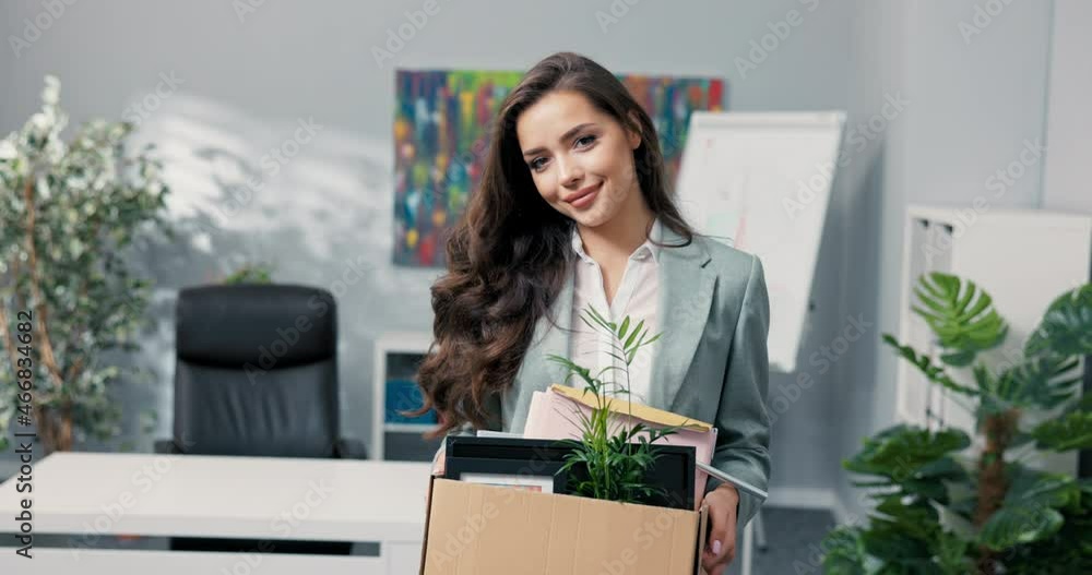 Attractive beautiful smiling brunette in shirt stands in front of desk in company office in hands holds box with packed things, promotion in position, coming to new job in office, employment, joy Attractive beautiful smiling brunette in shirt stands in front of desk in company office in hands holds box with packed things, promotion in position, coming to new job in office, employment, joy